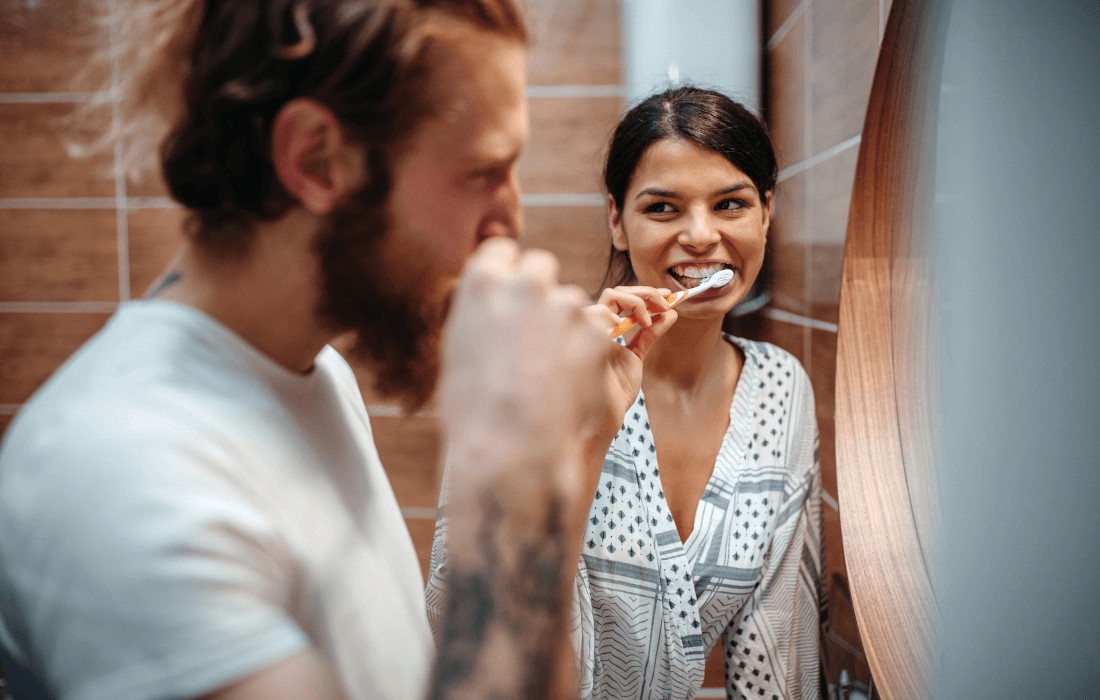 Ein Mann und eine Frau stehen gemeinsam im Bad, putzen sich die Zähne und lächeln sich dabei an.