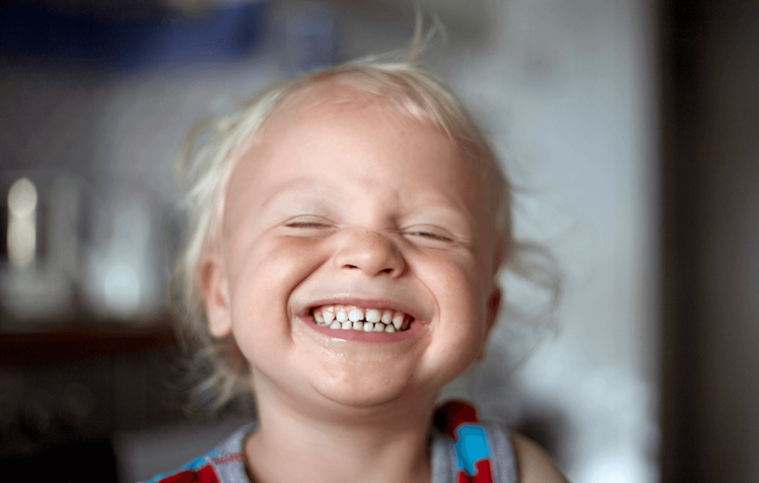 Ein Kleinkind mit weißblonden Haaren grinst über das ganze Gesicht und hält seine Milchzähne in die Kamera.